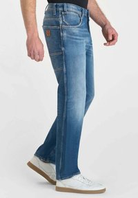 Jeans in denim blu con taglio dritto e finitura sbiadita. Caratteristiche includono cinque tasche, cuciture a vista e una patta in pelle sulla vita.