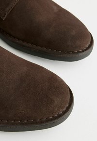 Zapatos de ante marrón con punta redondeada, costuras visibles y suela de goma negra. La textura parece suave y mate. Vista en primer plano.