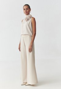 Top sans manches beige avec un col haut et des détails floraux, associé à un pantalon à jambes larges. Des chaussures pointues beige complètent le look.