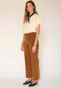 Haut à manches courtes crème, pantalon large en velours côtelé marron et ballerines noires avec des accents cloutés. Le mannequin porte une écharpe noire autour du cou.