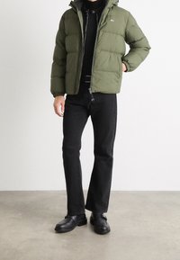 Vadderad, olivgrön jacka med dragkedja och huva, kombinerad med svarta denimbyxor och svarta skor, allt mot en ljus bakgrund.