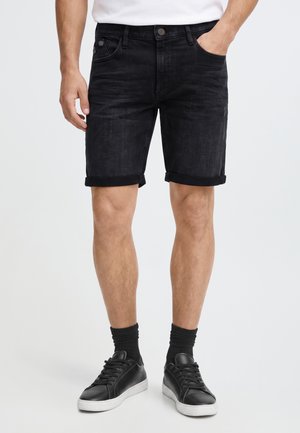 Un homme porte un short en jean noir roulé avec des chaussettes noires et des baskets noires, debout avec une main dans la poche sur un fond uni.
