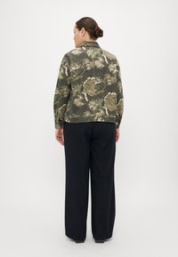 Veste à motif floral en vert et beige, avec un col et des accents de clous. Associée à un pantalon noir à jambes larges.