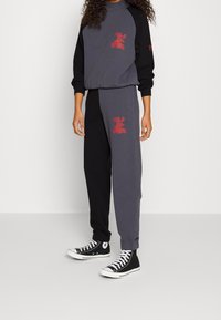 Mujer vistiendo un sudadera color bloque en negro y gris con logotipos rojos, combinada con joggers a juego y zapatillas altas negras.