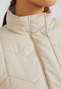 Giacca isolante beige chiaro con colletto alto, caratterizzata da un motivo trapuntato e una piccola tasca con zip sul davanti. Texture morbida.
