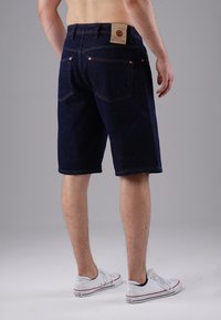 Denim shorts with a dark blue color, five-pocket design, straight fit, and branded patch on the back waistband. White sneakers complete the look.