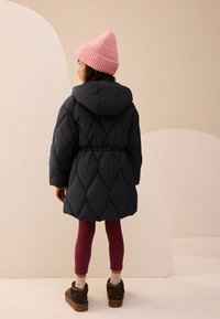 Veste matelassée noire à capuche avec taille cintrée, associée à des leggings bordeaux et des bottes marron à imprimé animalier, coiffée d'un bonnet rose.