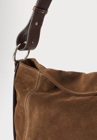 Bruine suède handtas met een zachte textuur en crème stiksels. Heeft een donkerleren schouderband en een metalen ring voor bevestiging.