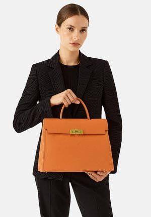 Orangene Lederhandtasche mit strukturiertem Design, oberen Traggriff und goldfarbenen Hardware-Akzenten; gehalten von einer Person in einem schwarzen gemusterten Blazer.