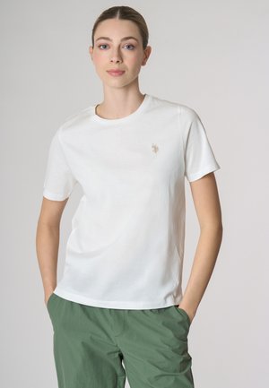 T-shirt bianco in cotone con maniche corte, collo rotondo, presenta un piccolo logo beige sul lato sinistro del petto. Abbinato a pantaloni verdi.