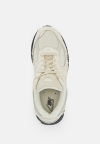 Beige sportcipő, hálós és velúrból készült, kerek fűzőkkel, párnázott nyelvvel és fekete gumitalppal, amely texturált mintákkal rendelkezik.