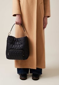 Black quilted hobo bag with a hexagonal pattern, featuring a front zip pocket and a short leather handle, held by a person in a camel coat.