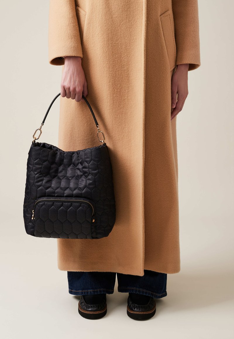 Black quilted hobo bag with a hexagonal pattern, featuring a front zip pocket and a short leather handle, held by a person in a camel coat.