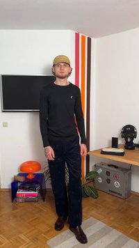 Fekete hosszú ujjú ing, sötétkék mintás nadrág, bézs sapka, sötétbarna cipő. Narancssárga és lila bútorok egy modern szobabelsőben.