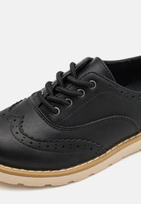 Scarpa nera in pelle con lacci, design con perforazioni e punta arrotondata, e suola in gomma beige. Presenta lacci neri e dettagli di cucitura.