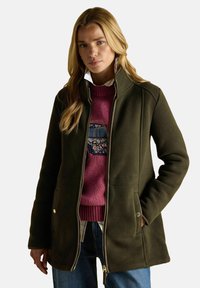 Blondynka w zielonej kurtce na zamek, nosząca różowy sweter z kwiatowym wzorem oraz niebieskie dżinsy, stoi z jedną ręką w kieszeni.