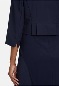 Námorně modrý peplum top se strukturovaným designem, s tříčtvrtečními rukávy a postranním rozparkem na lemu. Hladká, lehce texturovaná tkanina.