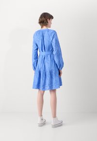 Rochie albastră cu mâneci lungi și fustă în straturi, decorată cu broderie florală, talie ajustată cu o curea și asortată cu adidași albi.