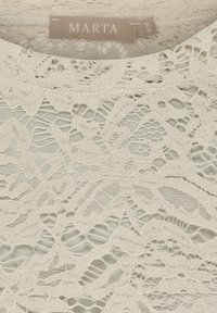 Elfenbensfarvet blonderstof med indviklede blomsterdekor, der har en tekstureret overflade og et blødt, delikat design. Mærket er 'MARTA' og størrelse 'S'.