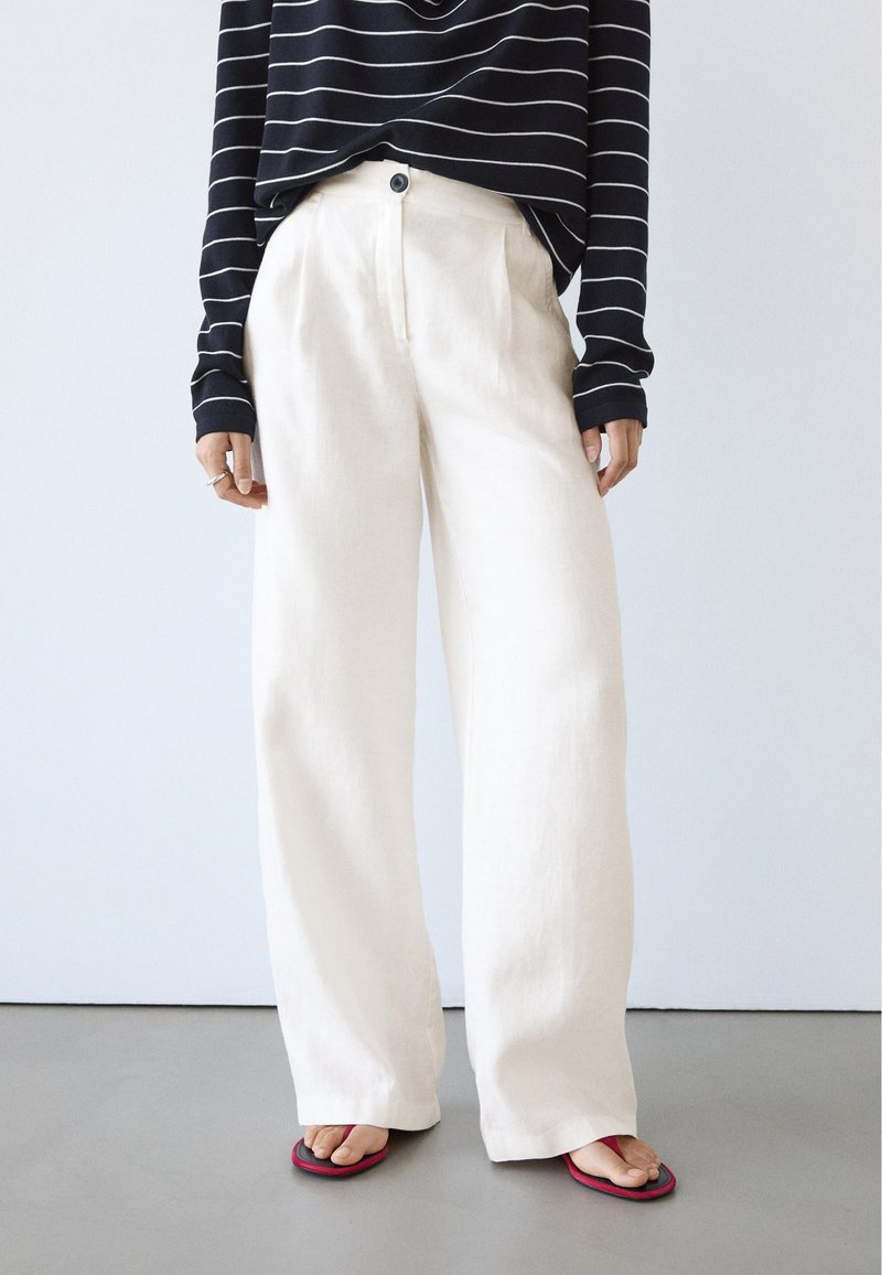 Personne portant un pantalon blanc ample, un haut à manches longues rayé noir et blanc, et des sandales rouges, debout sur un sol gris devant un mur blanc.