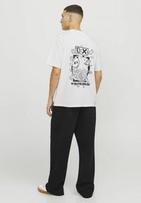 Hvid bomulds-t-shirt med et sort grafisk motiv af en sparegris omgivet af pengesedler på ryggen, parret med sorte løstsiddende bukser.