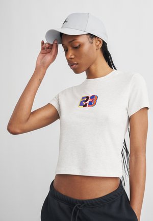 Young person adjusting light gray cap, wearing white cropped T-shirt with colorful "23" on front and black drawstring pants.