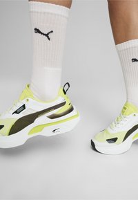 Scarpe sportive bianche e gialle con dettagli neri, in materiale mesh e sintetico, con suola spessa, abbinate a calze bianche a costine con un logo.
