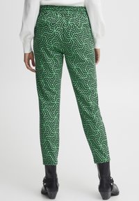 Grüne gemusterte Hose mit geometrischem schwarzem Design, elastischem Bund und schmal zulaufendem Bein, kombiniert mit schwarzen Knöchelstiefeln.
