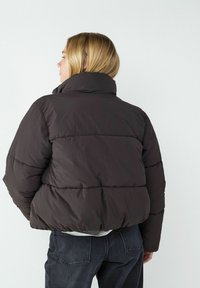 Braune Daunenjacke mit hohem Kragen und gestepptem Design. Sie hat einen cropped Schnitt und eine glatte Textur und wird mit dunklen Jeans kombiniert.