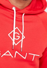 Sweat à capuche rouge avec cordons blancs et grand logo brodé en forme de losange. Tissu doux, avec un design de texte blanc audacieux.