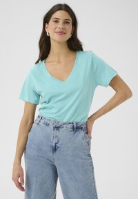 Tričko s výstřihem do V a kr átkým rukávem v světle tyrkysové bavlně. Nosí se s vysokým pasem a volným střihem v světle modrých džínách, které představují neformální outfit.