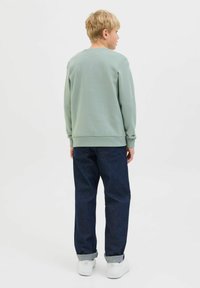 Sweatshirt vert à manches longues, col rond ; associé à un jean bleu foncé et des baskets blanches ; couture arrière et poignets visibles.