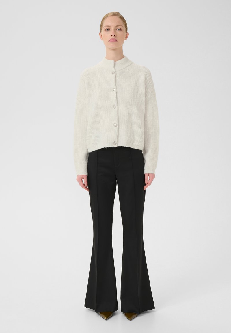 Cardigan blanc à boutons avec un col haut et un tissu texturé, associé à un pantalon noir évasé et des chaussures à bout pointu.
