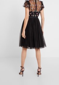 Robe en tulle noir avec broderies florales sur un corsage transparent, manches courtes à volants et jupe évasée ; associée à des escarpins à lanières.