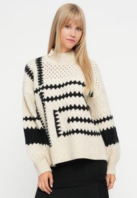 Cremefarvet og sort striktrøje med høj hals, der har geometriske mønstre og teksturerede detaljer. Løs pasform med ribbede kanter og manchetter.