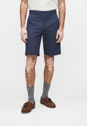 Άνδρας που φοράει ναυτικού μπλε σορτς, γκρι κάλτσες και καφέ δερμάτινα παπούτσια για σκάφος, στέκεται μπροστά σε ένα απλό ανοιχτό φόντο.