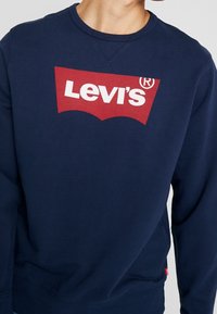 Marinblå sweatshirt i bomullblandning med en röd Levi's-logotyp och en klassisk rundhalsning. Mjuk textur med ribbade ärmslut.
