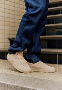 Zapatillas de ante beige con un perfil bajo, suelas planas y cordones. Combinadas con jeans de mezclilla azul oscuro, que muestran un corte relajado y con dobladillo.