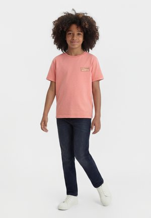 Kind met krullend haar, gekleed in een koraalrood t-shirt, donkerblauwe spijkerbroek en witte sneakers, staand tegen een eenvoudige lichte achtergrond.