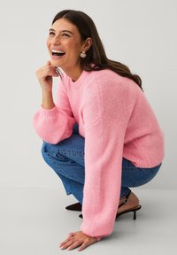 Maglione rosa lavorato a maglia con maniche a palloncino, texture morbida e scollo tondo, abbinato a jeans blu e sandali marroni con tacco basso.