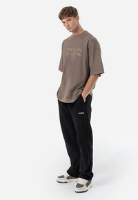 Didelis rudos spalvos trumpomis rankovėmis marškinėlis su reljefiniu logotipu, derinamas su juodomis sportinėmis kelnėmis su baltu prekės ženklu. Modelis stovėdamas su rankomis kišenėse.