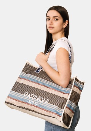 Junge Frau mit langem dunklem Haar, die ein weißes Hemd trägt und eine große gestreifte Tragetasche mit dem Schriftzug "Gattinoni Roma" bei sich hat.