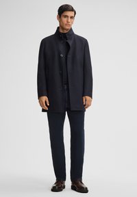 Marineblauwe wollen coat met hoge kraag, knopen aan de voorkant en zijzakken, gecombineerd met donkere jeans en bruine leren schoenen.