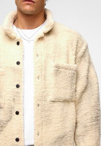 Lichtbeige fleece jack met een knoopsluiting aan de voorkant, een kraag en een patchzak. De stof heeft een zachte, gestructureerde uitstraling.