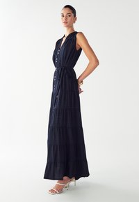 Námornícky modrá, bezrukávová maxi šaty s viacvrstvovým dizajnom, so zapínaním na gombíky vpredu, opaskom v páse a jemnou textúrou. Skombinované s bielymi sandálmi na opätku.