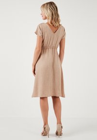 Beige ľanové šaty s krátkymi rukávmi a V-výstrihom. Obsahujú nariasený pás, dĺžku po kolená a hladkú štruktúru. Skombinované s béžovými podpätkami.