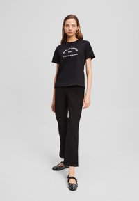 T-shirt nera di cotone con testo bianco, abbinata a pantaloni neri sartoriali. Scarpe nere con borchie bianche completano il look.
