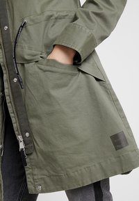 Olivově zelená bunda se zipovým zapínáním, bočními kapsami a cedulkou. Má vypasovaný pas a texturovanou látku s odolným povrchem.