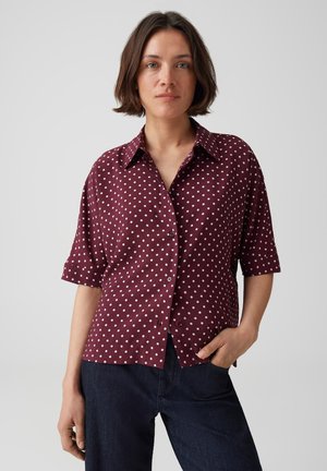 Vrouw met kort bruin haar, gekleed in een bordeauxrood kort mouwen overhemd met witte stippen en donkerblauwe jeans tegen een effen achtergrond.