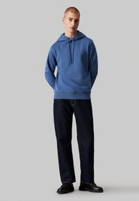 Felpa con cappuccio blu realizzata in tessuto morbido, dotata di coulisse, polsini a coste e un piccolo logo sul petto. Abbinata a jeans in denim scuro.